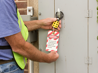 A person's hands are shown attaching a yellow lockout device with a red and white "DANGER" tag to a metal box or panel